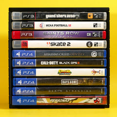 A black plastic game case organizer holding nine video game discs vertically, set against a solid bright yellow background. The discs belong to PlayStation 3 (PS3) and PlayStation 4 (PS4), including titles such as Grand Theft Auto, Saints Row, Skate 2, Assassin's Creed Odyssey, Call of Duty: Black Ops III, Crash Team Racing, Days Gone, Death Stranding, and Persona 5.